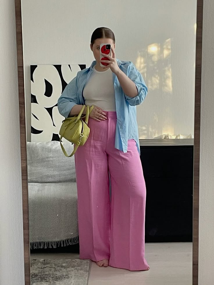 Woman in pastel spring outfit wearing a white tee, light blue oversized shirt, pink wide-leg pants, and holding a chartreuse handbag, taking a mirror selfie in a modern room.