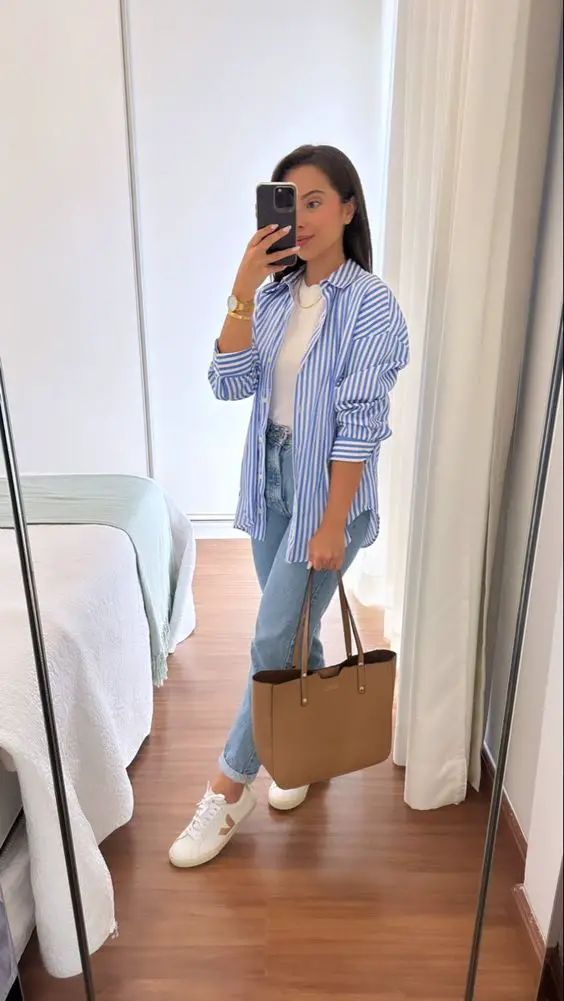 Casual striped shirt outfit with a woman wearing a blue-and-white button-down over a white tee, light-wash jeans, white sneakers, and a camel tote bag, posing indoors.