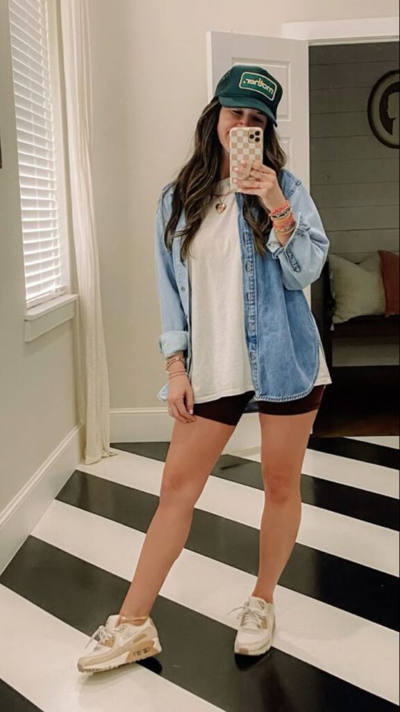 Sporty casual spring outfit with a green cap, oversized denim shirt, white tee, black biker shorts, and beige sneakers, styled with layered bracelets.