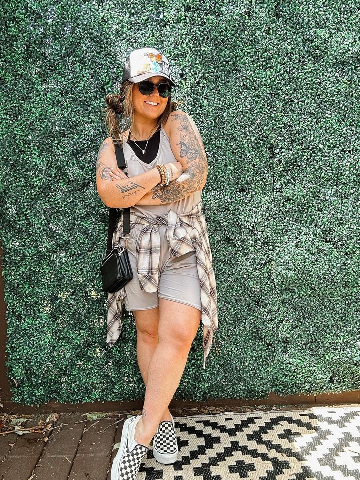 Trendy laid-back look with a taupe biker short set, plaid shirt tied around the waist, floral trucker hat, checkered Vans slip-ons, and crossbody bag.