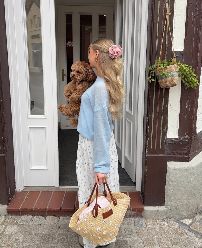 Romantic cottagecore outfit with a light blue sweater, white floral maxi skirt, straw tote with daisies, and a large pink flower hair clip, holding a small dog.