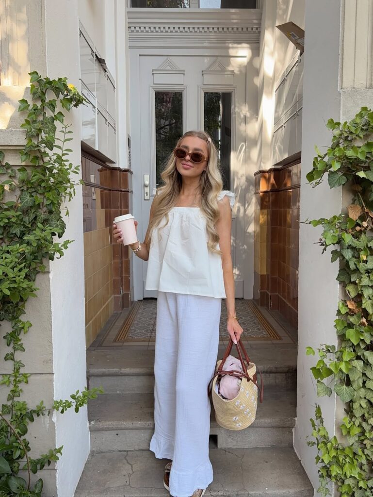Monochrome white flow outfit featuring a ruffle-strap white top, wide gauzy white pants, oversized sunglasses, and a straw tote with spring layers inside.

