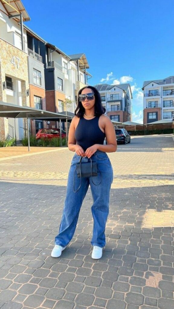 Woman wearing a black sleeveless tank, relaxed blue jeans, white sneakers, oversized sunglasses, and holding a tiny black handbag, standing in a sunlit modern apartment complex lot.