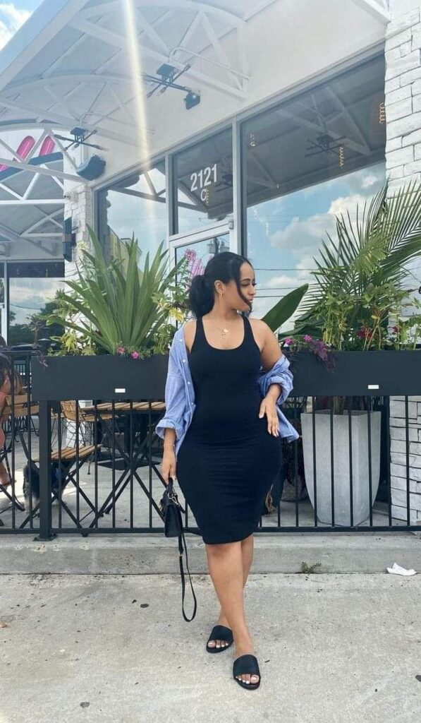 Woman wearing a fitted black tank dress with an open light blue button-up shirt, black slide sandals, and a small black handbag, standing confidently outside a modern café.