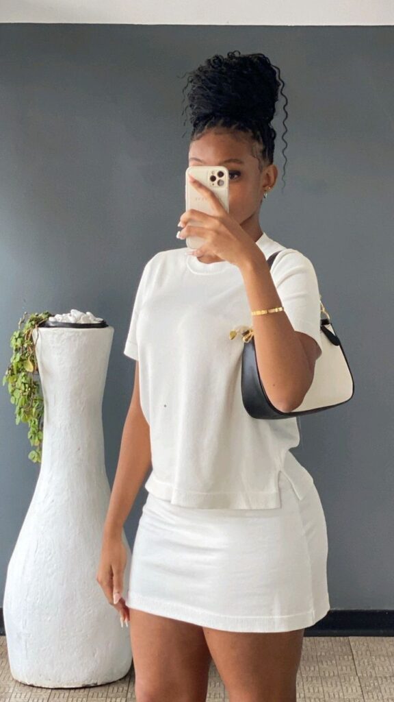 Woman taking a mirror selfie wearing a white short-sleeve top and fitted white mini skirt, styled with a black-and-white shoulder bag, gold bracelet, and high braided bun.