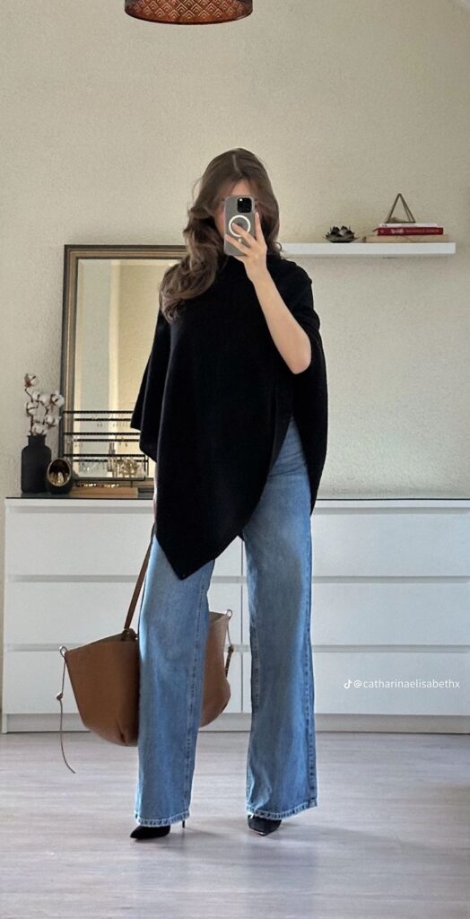 Woman wearing a black asymmetrical poncho top, light-wash wide-leg jeans, black pointed-toe shoes, and carrying a large brown tote, posing for a mirror selfie in a modern bedroom.