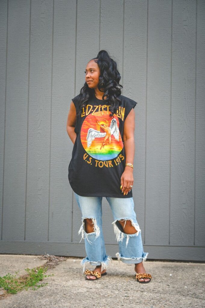 Woman wearing an oversized Led Zeppelin graphic tank top with heavily distressed ripped jeans and leopard print sandals, standing against a gray paneled wall with a casual, edgy vibe.
