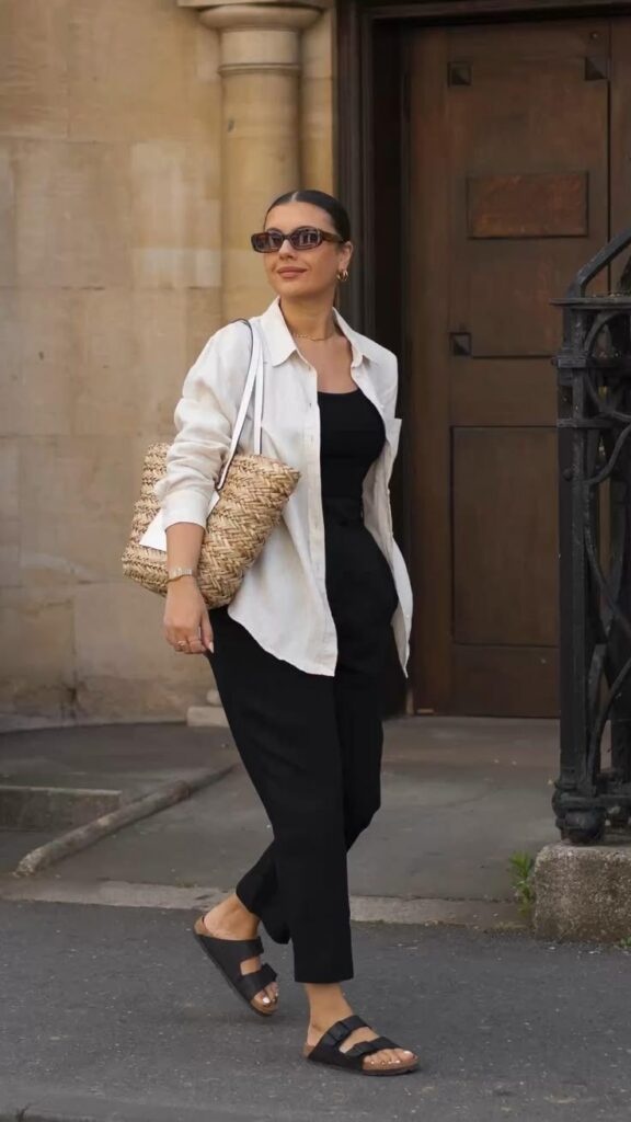 Woman wearing a relaxed, chic spring outfit with a black top and pants, a cream-colored button-down shirt, black sandals, and a woven tote bag. She’s walking confidently in an urban setting with sunglasses and pulled-back hair.