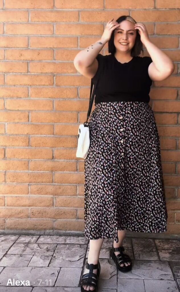 Woman wearing a black top and floral midi skirt, paired with black sandals and a white crossbody bag, standing against a brick wall and smiling confidently.