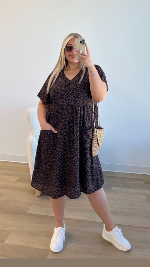 Woman wearing a flowy printed midi dress with white sneakers, a straw shoulder bag, and oversized sunglasses, taking a mirror selfie in a bright, modern room.