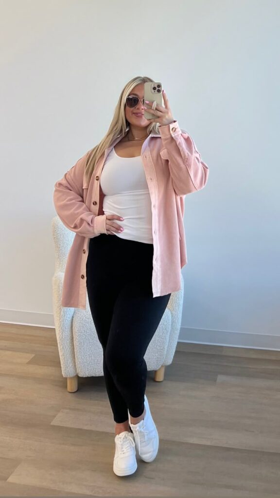 Woman wearing a pink button-up overshirt, white tank top, black leggings, and white sneakers, taking a mirror selfie in a minimal room with neutral tones.