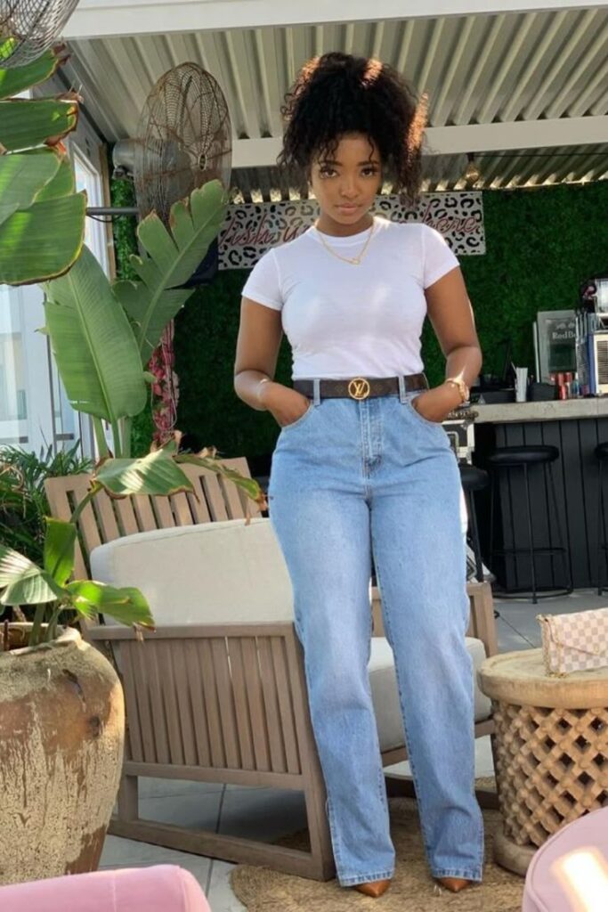 Woman wearing a fitted white t-shirt tucked into high-waisted light blue jeans, styled with a designer belt and gold jewelry, standing confidently in an outdoor lounge setting.