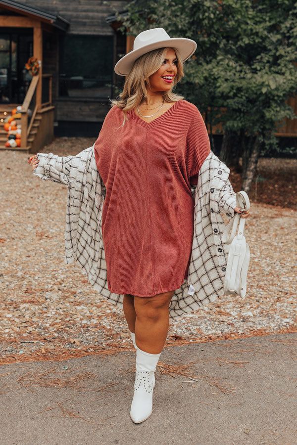 Woman wearing a rust sweater dress with a plaid shacket, white ankle boots, and a wide-brim hat, holding a white handbag and smiling outdoors in a rustic setting.