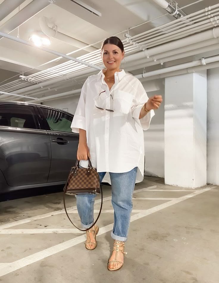 Woman wearing an oversized white button-up shirt with relaxed blue jeans, gold-studded flat sandals, and a brown checkered handbag, smiling in a modern parking garage.