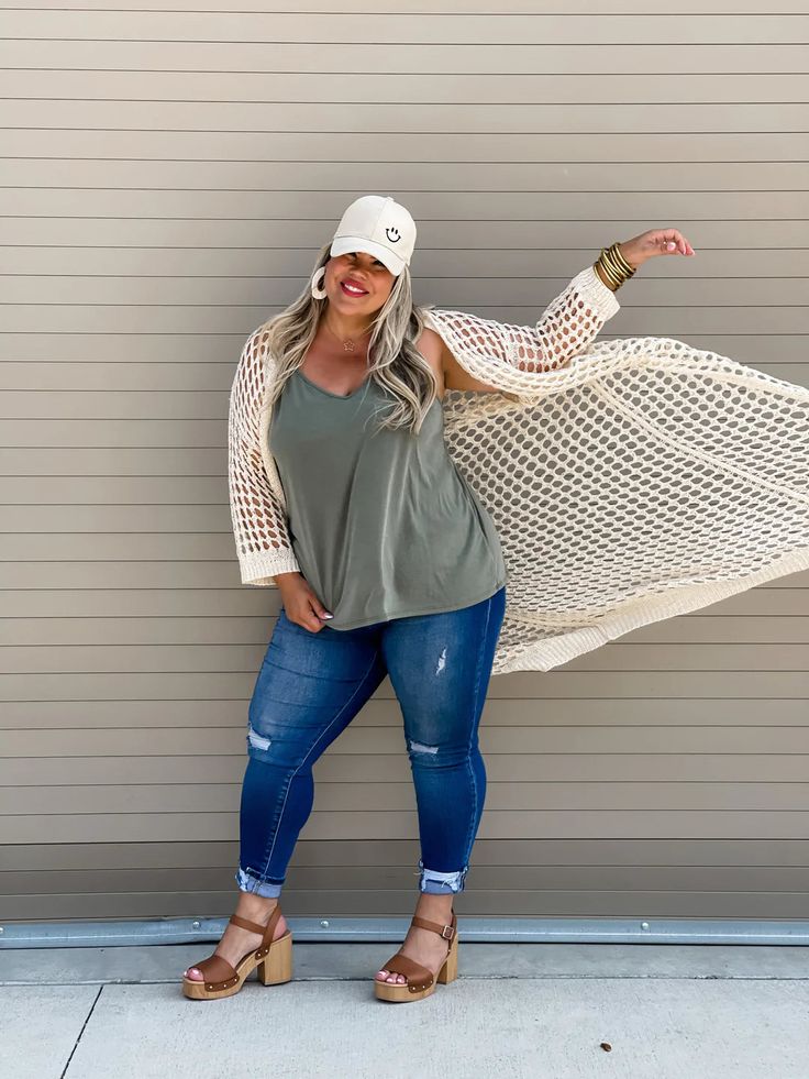 Woman wearing a loose olive tank, ripped skinny jeans, wooden platform sandals, a cream open-knit cardigan, and a smiley face cap, posing playfully against a tan wall.