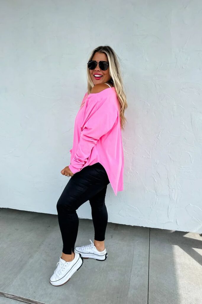 Woman wearing a hot pink oversized top, black leggings, and white platform sneakers with loose blonde hair and black sunglasses.