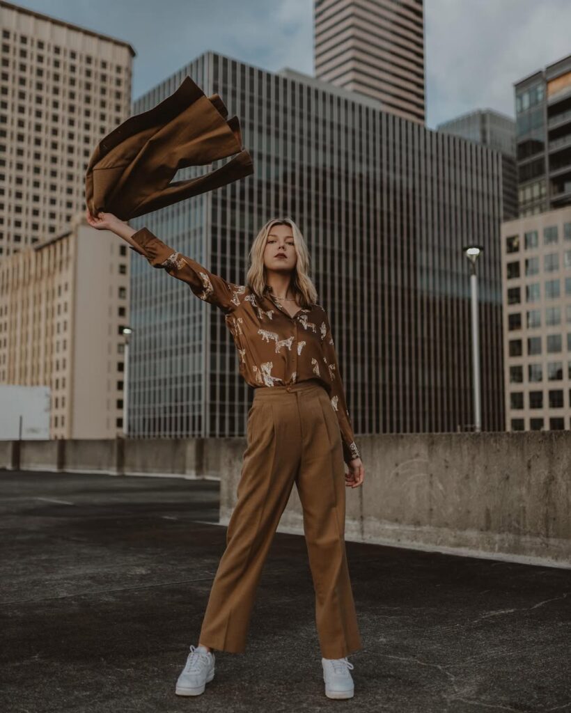 City rooftop outfit with a printed brown blouse tucked into tailored camel trousers, styled with white sneakers and a confident pose.