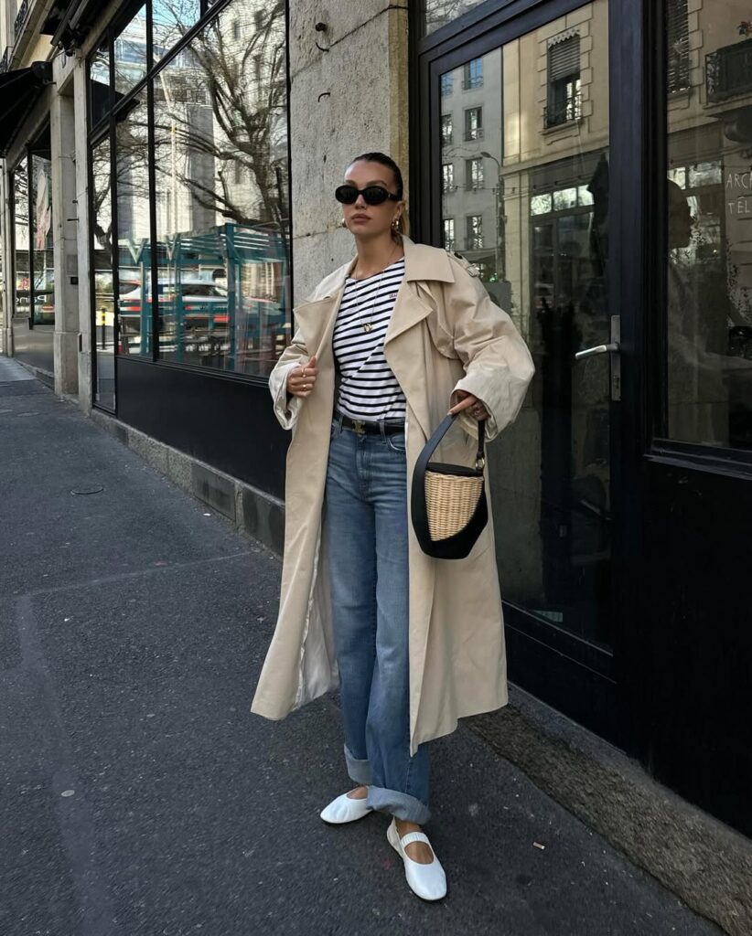 Parisian-inspired street style featuring a striped tee, beige trench coat, cuffed jeans, white ballet flats, and a straw-accented bag.