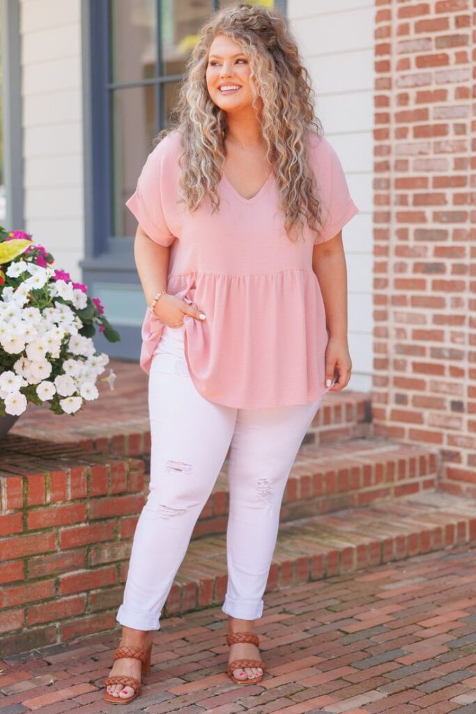 Plus-size woman in a blush pink peplum top, distressed white skinny jeans, and tan braided sandals, standing on a brick walkway.