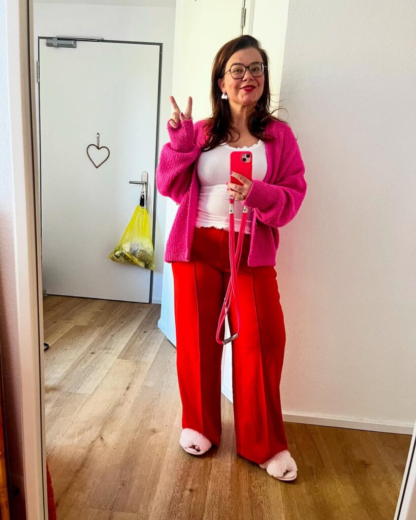 Woman in a bright pink cardigan, red wide-leg trousers, and a white ribbed tank top, taking a mirror selfie and flashing a peace sign.