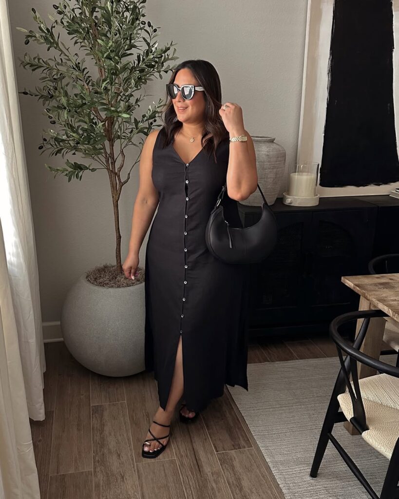Woman in a black sleeveless button-down maxi dress with a front slit, strappy black sandals, oversized sunglasses, and a curved black bag.