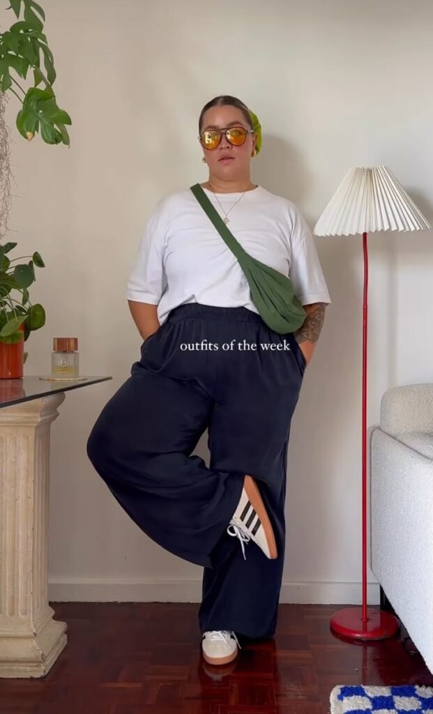Sporty-chic look with a white oversized T-shirt, loose navy wide-leg pants, Adidas sneakers, a green crossbody bag, and yellow-tinted sunglasses.
