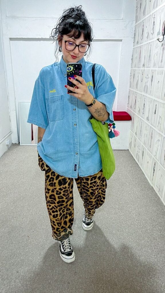 Eclectic street-style outfit with an oversized denim shirt, leopard print wide-leg pants, black Vans sneakers, and a lime green tote bag.