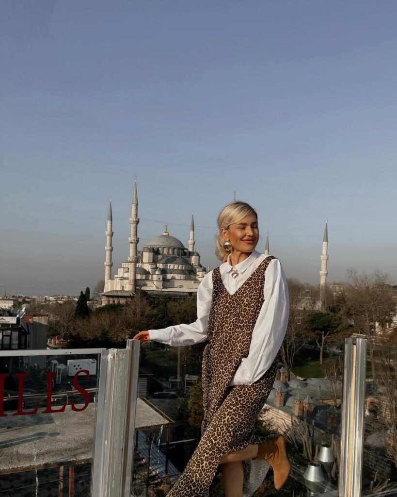 Stylish travel outfit idea for women over 40 featuring leopard slip dress layered over white shirt, tan suede ankle boots, and statement earrings with a mosque background.
