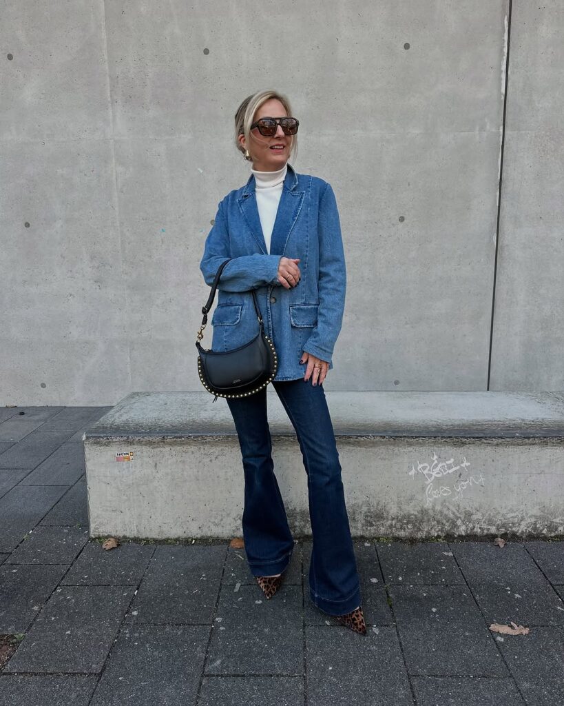 Stylish outfit idea for women over 40 featuring denim blazer, white turtleneck, dark flared jeans, leopard-print shoes, black studded handbag, and oversized sunglasses.

