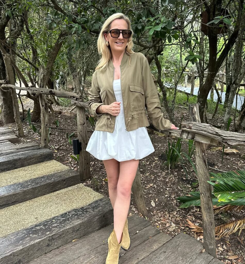 Casual chic outfit idea for women over 40 featuring khaki utility jacket, white mini dress, suede ankle boots, and oversized sunglasses in a natural outdoor setting.