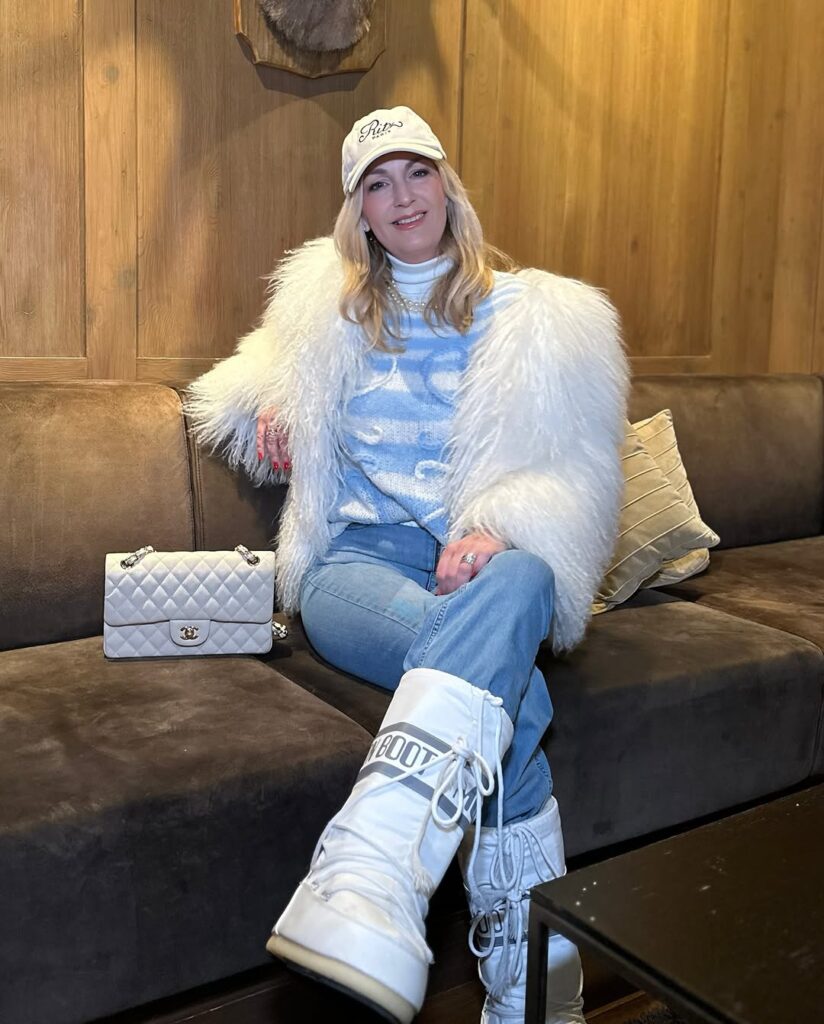 Cozy chic winter outfit idea for women over 40 featuring white faux fur jacket, blue patterned sweater, light-wash jeans, white Moon Boots, embroidered cap, and Chanel quilted handbag in a lodge setting.