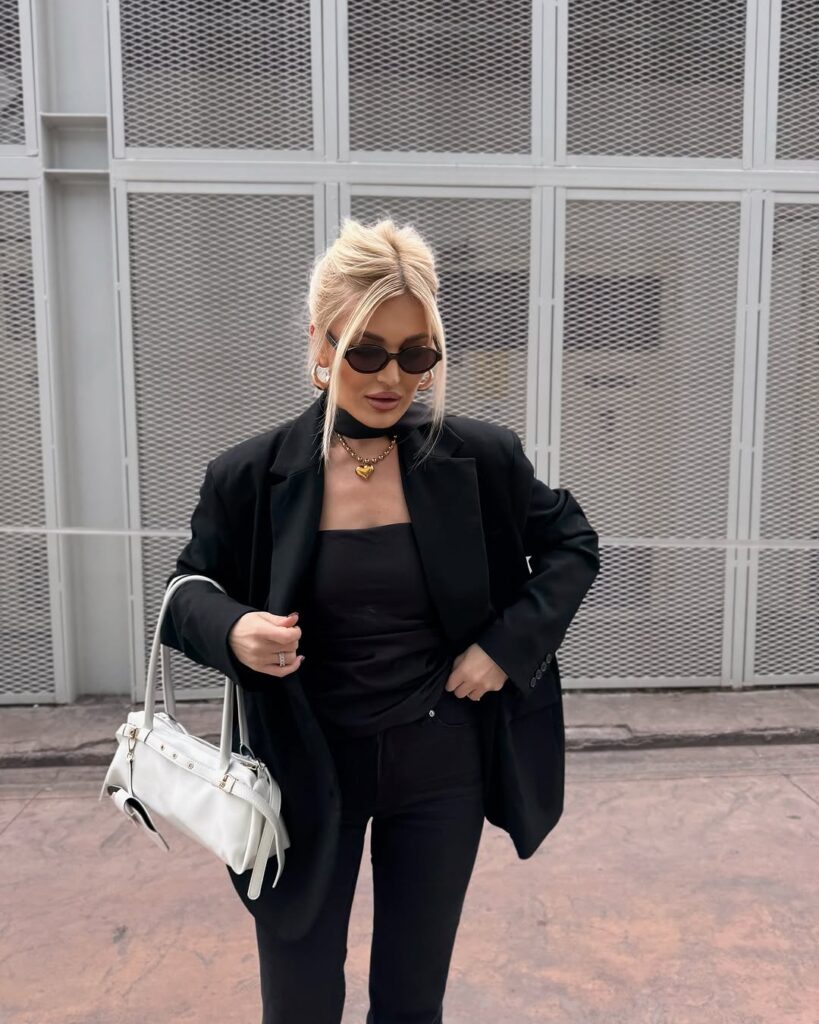 Chic all-black outfit idea for women over 40 featuring oversized blazer, strapless top, black jeans, gold heart necklace, hoop earrings, white handbag, and black sunglasses.