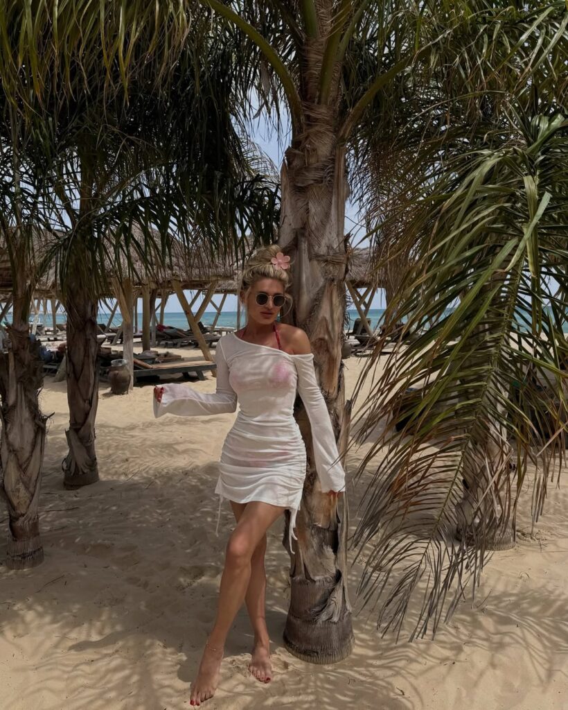Woman in a sheer white off-the-shoulder mini dress with a pink bikini underneath, barefoot on a tropical beach under palm trees.