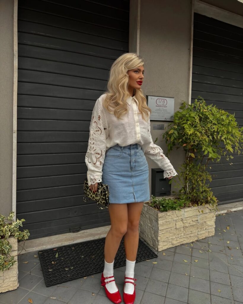 Woman wearing a white eyelet blouse, denim mini skirt, white socks with red Mary Jane shoes, holding a leopard clutch, standing on a city sidewalk.