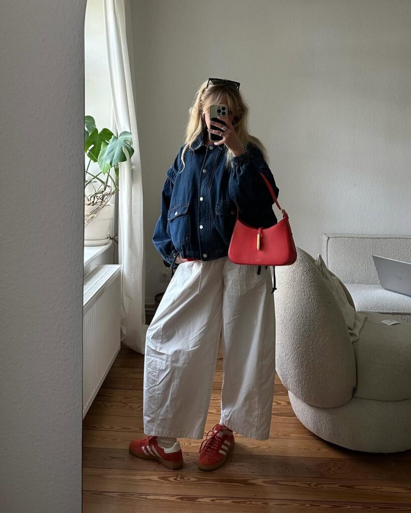 Woman taking a mirror selfie wearing an oversized dark denim jacket, wide-leg white pants, red sneakers, and a red shoulder bag indoors.