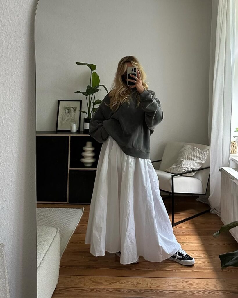Woman taking a mirror selfie wearing a charcoal oversized sweatshirt, white voluminous maxi skirt, and black low-top sneakers in a minimalist living room.