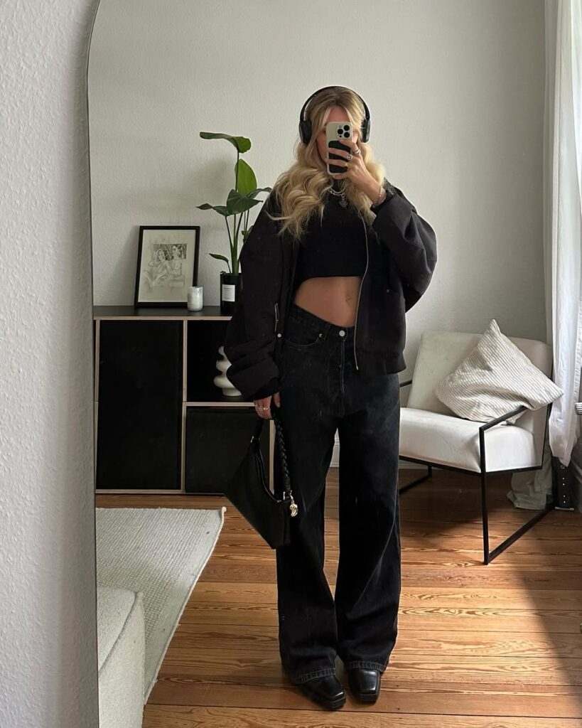 Woman taking a mirror selfie wearing a cropped black top, oversized black jeans, a black bomber jacket, headphones, and square-toe boots, holding a small black handbag.