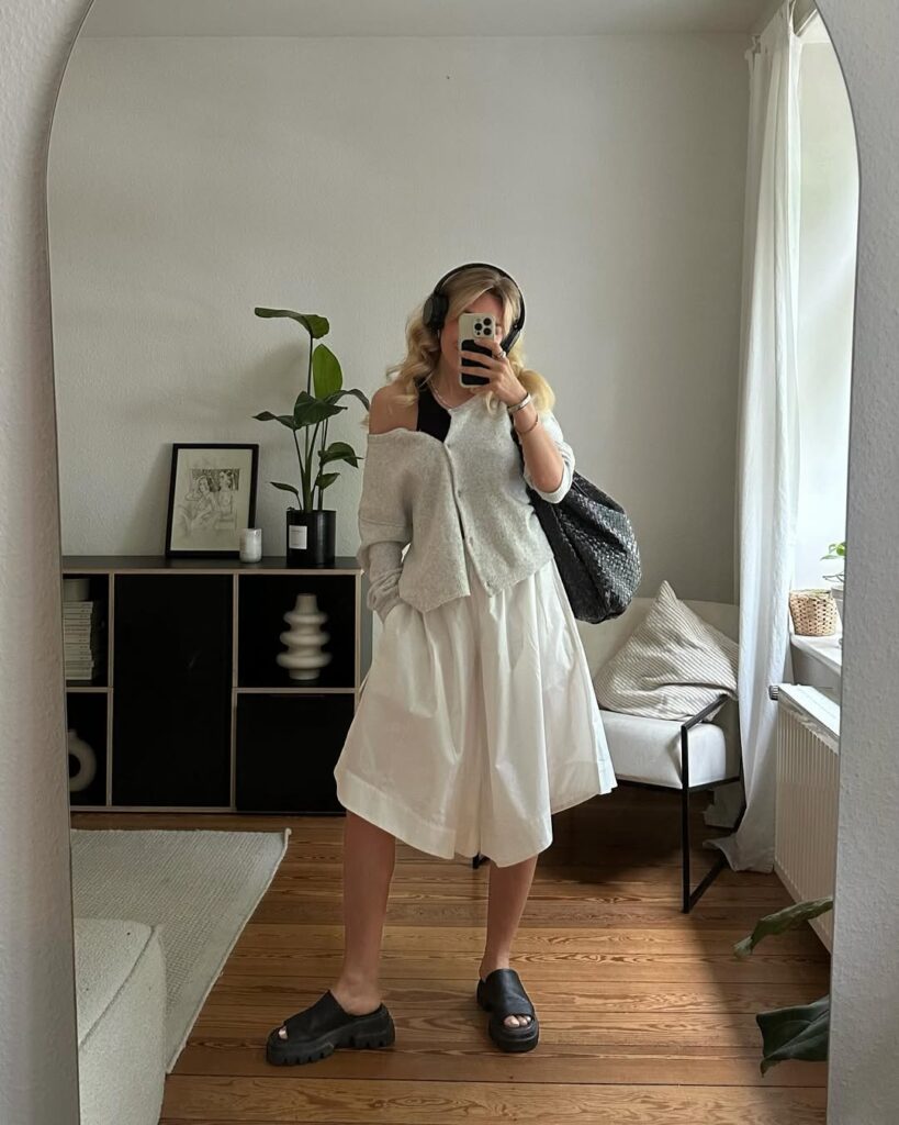 Woman taking a mirror selfie wearing a grey cardigan, black tank top, voluminous white culottes, black chunky slides, and headphones, carrying a slouchy black bag.