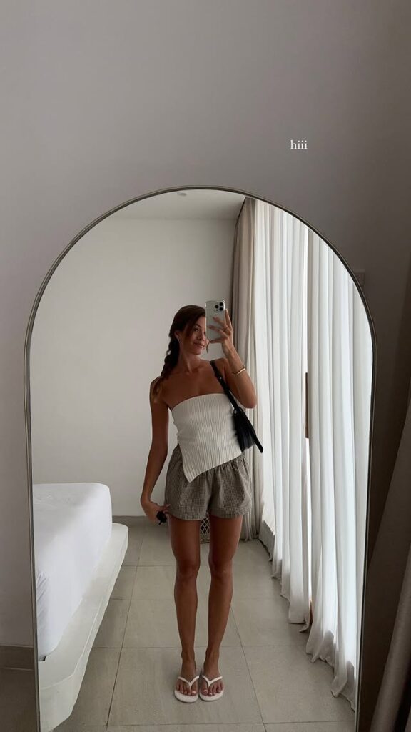 Woman taking a mirror selfie wearing a white strapless asymmetric top, gingham shorts, white flip-flops, and a black shoulder bag in a minimal, sunlit room.
