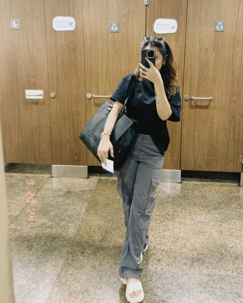 Woman taking a mirror selfie wearing an oversized black t-shirt, gray cargo pants, white slides, and carrying a large black tote bag in a public restroom.