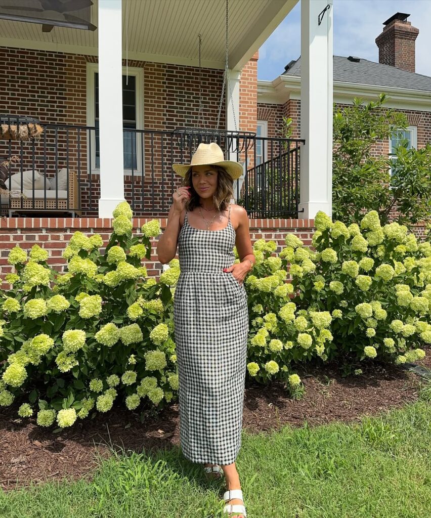 Woman wearing a black-and-white gingham sundress, straw hat, and white sandals, standing in front of blooming hydrangeas and a brick porch.