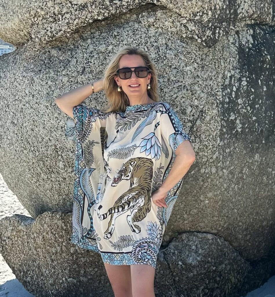 Woman wearing a printed tiger and botanical kaftan with oversized sunglasses, posing against large beach rocks.