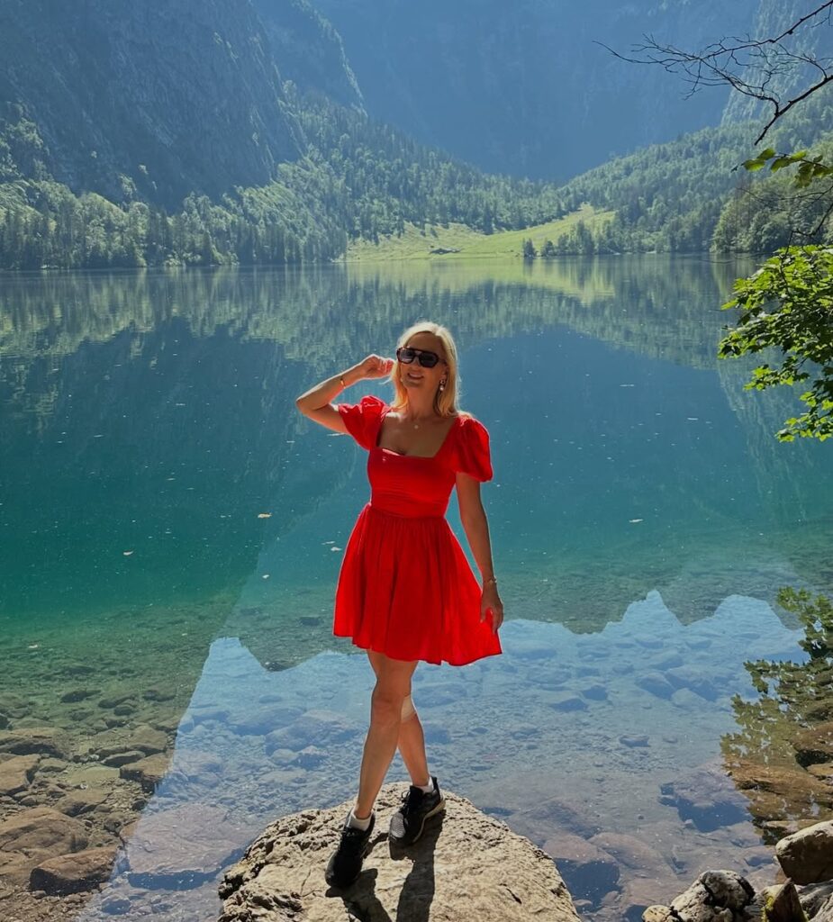 Woman in a bright red puff-sleeve mini dress and black sneakers standing on a rock by a clear, reflective mountain lake.