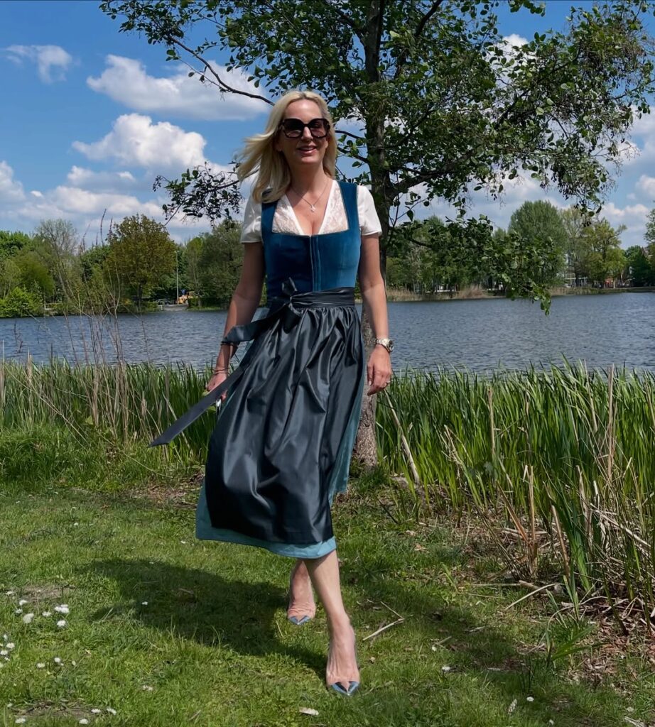 Woman wearing a teal dirndl dress with a black apron, lace blouse, and pointed-toe heels, walking near a lakeside on a sunny day.